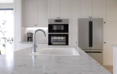 Ada ve gerekli tüm aletlerle dolu büyük beyaz bir mutfak. 3D görüntüleme. Üzerinde beyaz mermer olan bir taş tepeye vurgu, arka planda bir fırın, mikserden bir lavabo ve bir buzdolabı var..