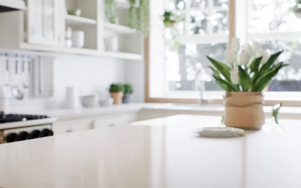 Close-up of a marble countertop of an island kitchen against a blurred background of a kitchen with appliances and utensils by a window with a green plant. 3d rendering