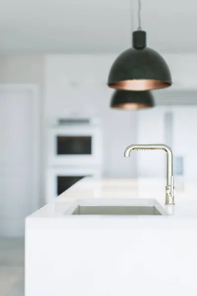 An image of a white kitchen with appliances, light fixtures and a view of the sink and faucet. The background is blurred, the focus is on the kitchen faucet. 3D rendering.