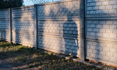 Düşük açılı güneş ışığıyla aydınlatılan tuğla gibi desenli sağlam beton bir çit yüzeyine ve çimenli zemine uzanan belirgin gölgeleri gösteriyor..