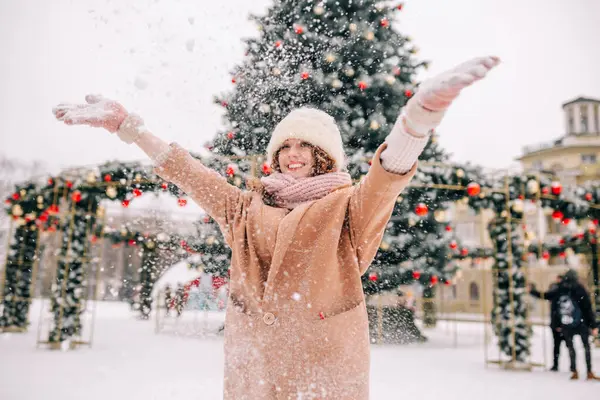 A curly girl blows snow against the backdrop of a beautiful New Year tree on a beautiful winter day. A woman plays in the snow in December