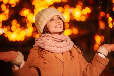 Akşamları büyüleyici kıvırcık saçlı genç bir bayan. Işıklar ve süslü bir Noel ağacı çerçeveli. Noel arifesinde şenlikli şehirde gece gezintisi