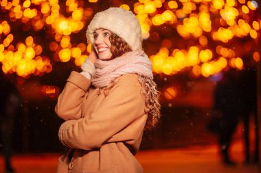 Kıvırcık saçlı, örgü şapkalı ve eşarplı genç bir kadın Noel ışıkları ve hoş bir kış akşamında şenlikli dekorasyonlar arasında poz veriyor.