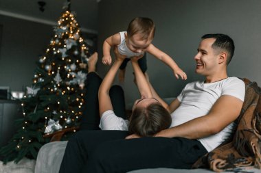 Anne, baba ve küçük çocuk mutlu bir şekilde evde sıcak ve rahat bir atmosferde yeni yılı kutluyorlar. Mutlu aile oyunları Noel ağacının yanında. Kış tatili ve insanların konsepti