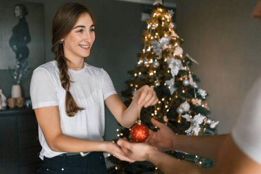 Genç ve güzel bir kadın Noel arifesinde Noel ağacını süslerle süslüyor. Şenlikli, rahat ev atmosferi kavramı. Kız gülümsüyor ve elinde bir Noel süsü tutuyor ağacın arka planına ve ışıklara karşı.