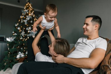 Anne, baba ve küçük çocuk mutlu bir şekilde evde sıcak ve rahat bir atmosferde yeni yılı kutluyorlar. Mutlu aile oyunları Noel ağacının yanında. Kış tatili ve insanların konsepti