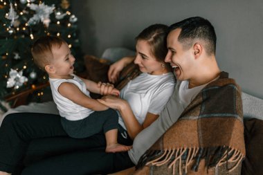 Mutlu aile oyunları Noel ağacının yanında. Anne, baba ve küçük çocuk mutlu bir şekilde yeni yıl tatilini evde rahat bir ortamda geçiriyorlar.