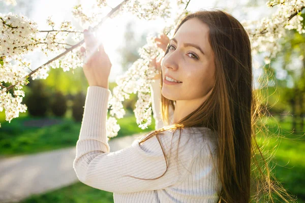 Gün batımında ilkbaharın erken saatlerinde çiçek açan bir elma ağacının yanında poz veren dişleri olan çekici bir kadının portresi.