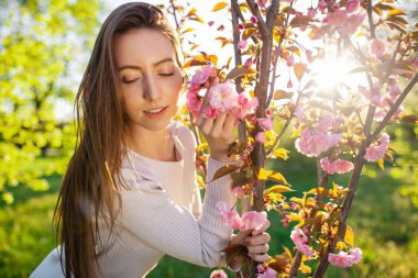 Güzel bir bahar akşamında, pembe çiçekli elma kiraz ağacının yanında poz veren, dalgalı saçlı büyüleyici bir kadın.