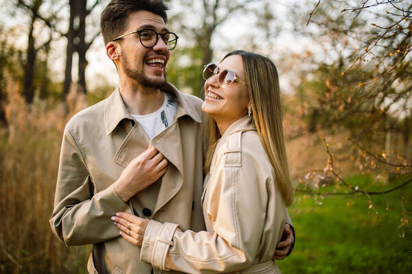 A man looks at a woman with love and tenderness. Close shot of a beautiful young couple in glasses and beige trench coats posing against the backdrop of a spring forest park