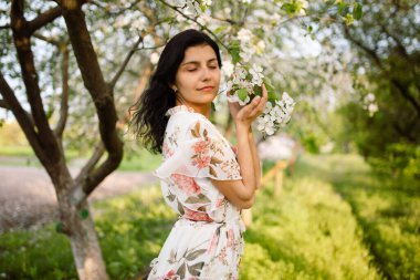 Parktaki bir bahçede çiçek desenli bir elbise giymiş, çiçek açan bir kiraz ağacının yanında poz veren çekici bir esmerin yakın plan portresi. Çıplak makyaj malzemesi. Portreyi kapat