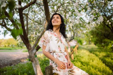 Mutlu genç bir kadın arka planda ağaçların açtığı bir bahar bahçesi parkında bir bankta oturuyor. Bahar geldi.