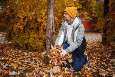 Eldivenli bir adam yere çömelmiş, düşen sonbahar yapraklarını topluyor. Turuncu, kırmızı ve sarı tonlarda renkli yapraklarla çevrili, mevsimin huzurlu özünü seviyor.