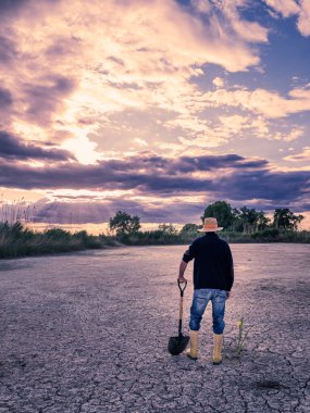 Kuru arazi için lastik botlarla. Çiftçi lastik çizmeleri ve küreğiyle çorak bir tarlaya gider. Arkadan bak. Kuraklık kavramı.