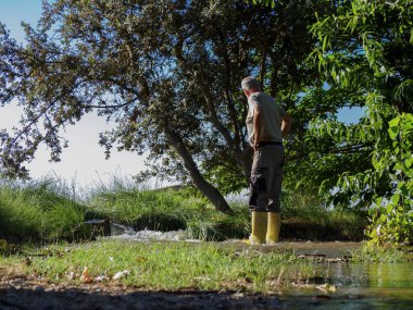 Botları olan yaşlı bir adam meyve ağaçlarını sulamak için suyu kanalize ediyor, tarlayı sel basıyor..