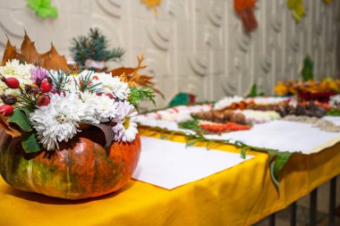 Canlı bir masa vitrininde renkli çiçekler ve sonbahar yapraklarıyla dolu bir balkabağı sergileniyor. Balkabağını çevreleyen çeşitli dekoratif öğeler sonbahar mevsimini kutluyor..