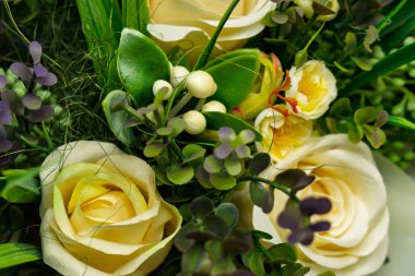 Zarif yeşil yapraklar ve narin beyaz aksanlarla sarmalanmış yumuşak sarı güller içeren dekoratif bir buketin ayrıntılı görüntüsü çiçek sanatını gözler önüne seriyor..