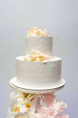 Güzel dekore edilmiş iki katlı bir düğün pastasında beyaz krema ve karmaşık çiçek aksanları bulunur. Basit bir stantta sergileniyor, özel bir kutlama için zarafetini artırıyor..