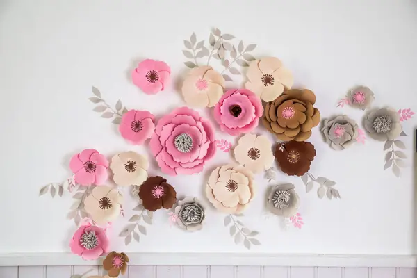 Beyaz bir duvarda sanatsal olarak canlı bir kağıt çiçek sergisi düzenlenir. Çiçekler çeşitli renk ve boyutlarda, odanın dekorunu taze ve yaratıcı bir dokunuşla genişletiyor..