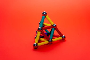 A three-dimensional triangle structure crafted from vibrant magnetic building sticks and spheres rests on a bright orange background. The design showcases creativity and engineering principles. clipart