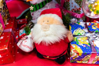 Şirin, pelüş bir Noel Baba, parlak hediyelerin arasına yerleşmiş, neşeli renkler ve desenler sergiliyor, tatil sezonunun sıcak ruhunu yakalıyor..