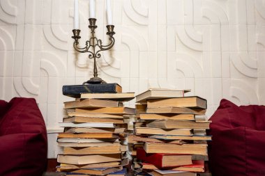 Bir yığın kitap bir mum tutacağının yanındaki masanın üzerinde duruyor. Arka plandaki yumuşak kırmızı yastıklar okuma ve rahatlama için sıcak ve davetkar bir atmosfer yaratıyor..