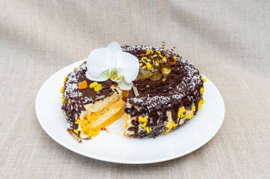 Çok güzel hazırlanmış bir çikolatalı pasta kremalı dolgu tabakalarına sahiptir ve sarı süslemeler ve beyaz bir orkideyle süslenir. Tarafsız bir arkaplanda, beyaz bir tabakta duruyor..