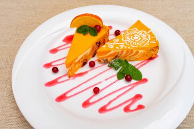 Çok güzel sunulmuş bir tatlıda, dekoratif bir sos üzerinde naneli yapraklar ve böğürtlenlerle süslenmiş iki dilim portakallı pasta yer alıyor. Neşeli bir kutlama için mükemmel..