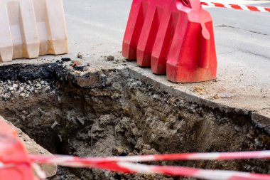 İşçiler yollarda güvenlik bariyerleriyle çevrili derin bir çukur kazıyorlar. Kırmızı ve beyaz bant, yoldan geçenlerin tamirat sırasında güvende olmalarını sağlayan tehlikeli bir bölgeyi işaret ediyor..