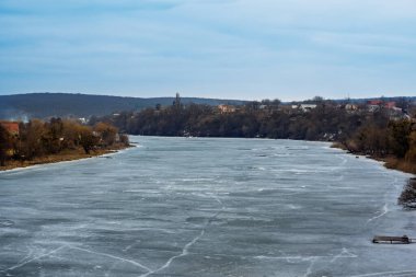 Sakin bir nehir, ağaçlar ve uzak binalarla çevrili buzla kaplıdır. Bulutlu gökyüzü, bu soğuk kış gününün sakin atmosferini yansıtıyor. Doğanın durgunluğunun güzelliğini yakalıyor..