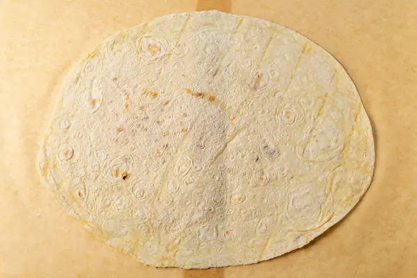 Büyük, yuvarlak bir tortilla bir parşömen kağıdının üzerinde duruyor. Hafifçe kahverengileşmiş yüzeyinde yemek pişirme, çeşitli lezzetli yemekler hazırlama belirtileri var..
