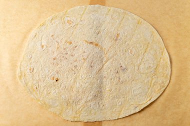 Büyük, yuvarlak bir tortilla bir parşömen kağıdının üzerinde duruyor. Hafifçe kahverengileşmiş yüzeyinde yemek pişirme, çeşitli lezzetli yemekler hazırlama belirtileri var..