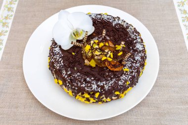 Güzel dekore edilmiş bir çikolatalı pasta, üzerinde orkide ve meyve olan beyaz bir tabakta oturur. Pastada çikolata parçacıkları ve parlak sarı süslemeler var. Kutlamalar için mükemmel..