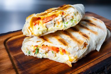 Tavuk, peynir, domates ve otlarla dolu nefis bir ızgara sargı sıcak bir mutfak ortamında ahşap bir kesme tahtasının üzerinde duruyor. Doku davetkar ve lezzetli..