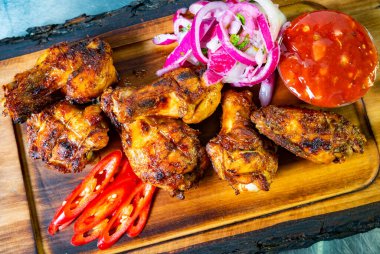 Lezzetli ızgara tavuk kanatları ahşap bir tabakta, yanında taze kırmızı soğan, baharatlı kırmızı salsa ve dilimlenmiş kırmızı biber ile birlikte düzenlenir. Canlı renkler yemeğin cazibesini arttırır.