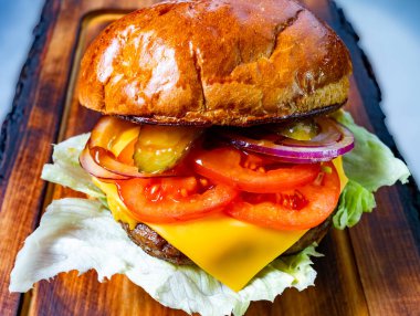 Lezzetli bir hamburgerin içinde erimiş peynir, taze domates, turşu, soğan ve kırsal ahşap bir tahtanın üzerinde sunulan kıtır marul katmanları yer alır..