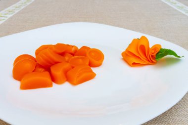 Taze turuncu havuç dilimleri, ince kesilmiş ve yeşil yaprakla süslenmiş dekoratif bir havuç çiçeğinin sergilendiği el değmemiş beyaz bir tabağa düzgünce yerleştirilir..