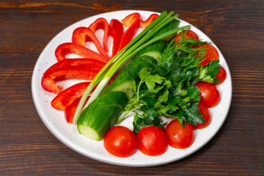 Taze sebzelerden oluşan renkli bir sergi kırmızı dolma biber, kiraz domatesleri, salatalık, yeşil soğan ve beyaz tabakta otlar. Canlı bir salata ya da garnitür için ideal..
