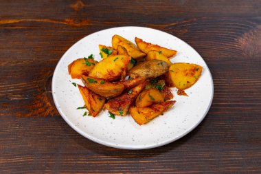 Koyu ahşap bir masanın üzerine yerleştirilmiş beyaz bir tabakta çıtır çıtır soslu altın sotelenmiş patatesler sunulur. Taze otlar, yemeğin cazibesini arttırmak için canlı renkler katar..