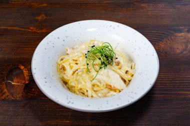Lezzetli kremalı bir makarna taze otlarla süslenmiş beyaz bir kasede sunulur. Tabak zengin bir sos içeriyor ve sıradan bir yemek deneyimini davet eden ahşap bir arka plana karşı hazırlanıyor..