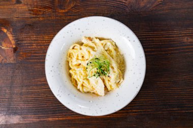 Bir tabak kremalı makarna dilimlenmiş tavuk ve narin yeşil otlarla kaplıdır. Tabak, benekli beyaz bir plaka üzerinde kırsal bir ahşap arka plana karşı servis edilir..