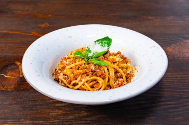 Bir tabak spagetti, ekmek kırıntıları ve taze otlarla kaplı beyaz bir kasede sunulur. Tabak kırsal ahşap bir masa üzerine kurulmuş, davetkar renkler ve dokular sergileniyor..