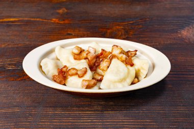 Sıcak hamur köfteleri krem rengi bir tabakta servis edilir, üzerinde altın, çıtır domuz parçaları vardır. Zengin, rahatlatıcı yemek kırsal bir ahşap masada duruyor, açlığı ve zevki davet ediyor..