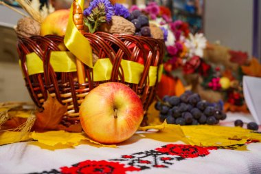 Elma, üzüm ve dekoratif elementlerle dolu dokuma sepet, sonbahar yapraklarıyla süslenmiş canlı bir masa örtüsünün üzerinde duruyor ve hasat mevsiminin bolluğunu kutluyor..