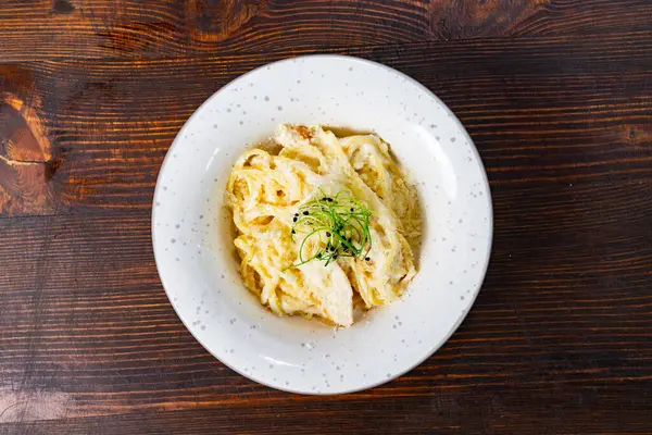 Bir tabak kremalı makarna dilimlenmiş tavuk ve narin yeşil otlarla kaplıdır. Tabak, benekli beyaz bir plaka üzerinde kırsal bir ahşap arka plana karşı servis edilir..