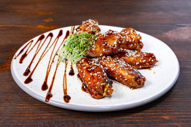 Lezzetli ızgara tavuk kanatları yuvarlak bir tabakta, susam tohumları ve soğanlarla süslenmiş, bol soslu, ahşap bir arka planda sergileniyor..