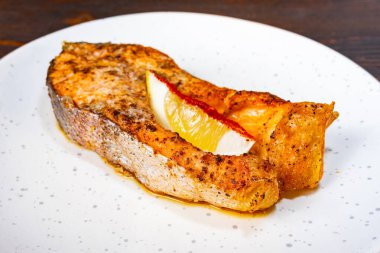 Güzel bir ızgara somon bifteği taze bir limon dilimi ile aksanlı, temiz beyaz bir tabakta duruyor. Balıklar zengin, çıtır çıtır bir dokuya ve iştah açıcı bir baharata sahiptir..