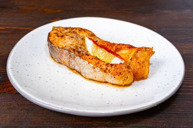 Bir dilim limonla süslenmiş güzel ızgara somon filetosu basit beyaz bir tabakta duruyor. Çanak, koyu ahşap bir zemine dayanıyor ve sıcak, davetkar bir atmosfer yaratıyor..