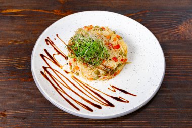 Düzgün düzenlenmiş bir kızarmış pirinç servisi üzerinde canlı sebzeler ve mikroyeşillikler barındırır. Tabak desenli beyaz bir tabağa yerleştiriliyor, koyu ahşap bir masa arkasına kuruluyor..