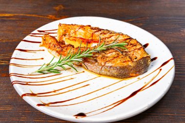 A perfectly grilled salmon steak rests on a white plate, garnished with fresh rosemary and drizzled with a rich sauce. The warm colors evoke a cozy dining atmosphere. clipart